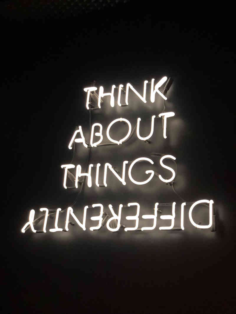Neon sign saying 'THINK ABOUT THINGS DIFFERENTLY', with all black background.  It has one letter per line, and 'DIFFERENTLY' is reversed